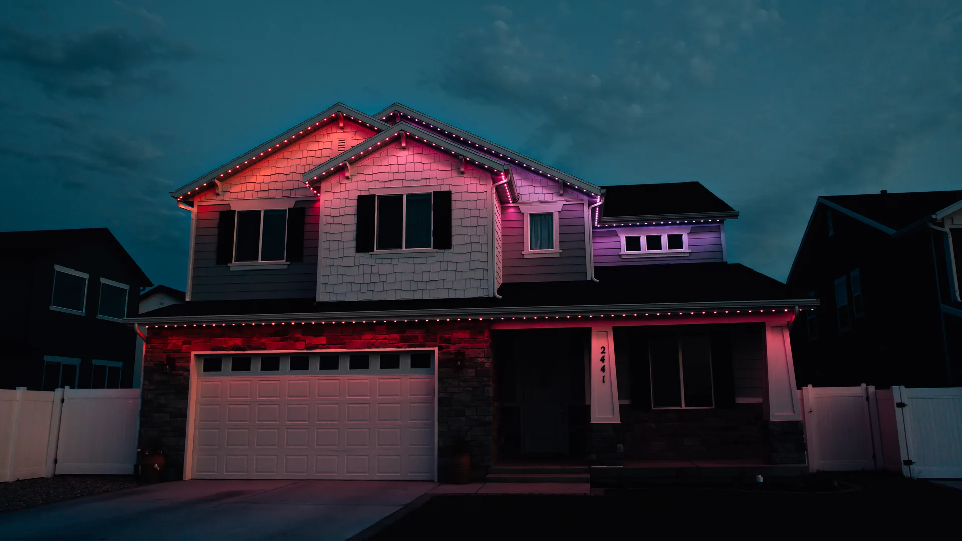 Two-story farmhouse at night with permanent architectural lighting glowing purple and teal along the roofline and porch