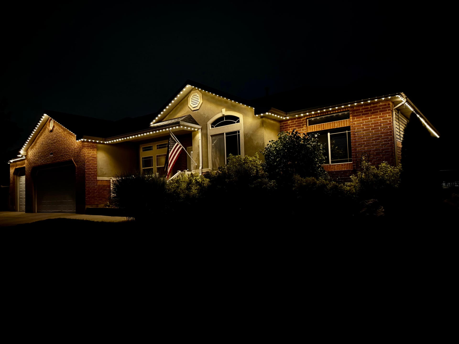 Brick and stucco home with warm white roofline lighting and an American flag