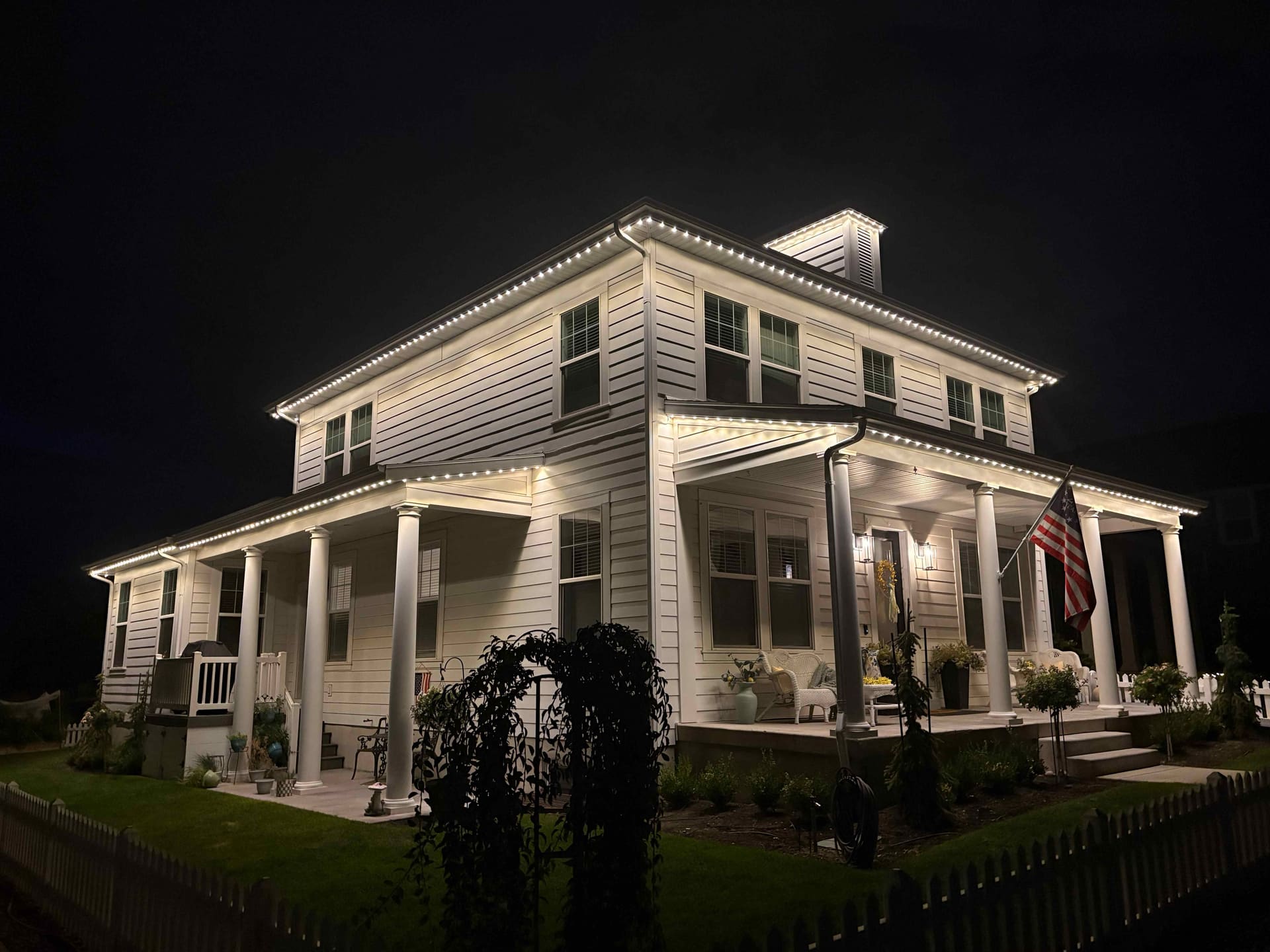 Large white farmhouse with warm white roofline lighting and a wraparound porch