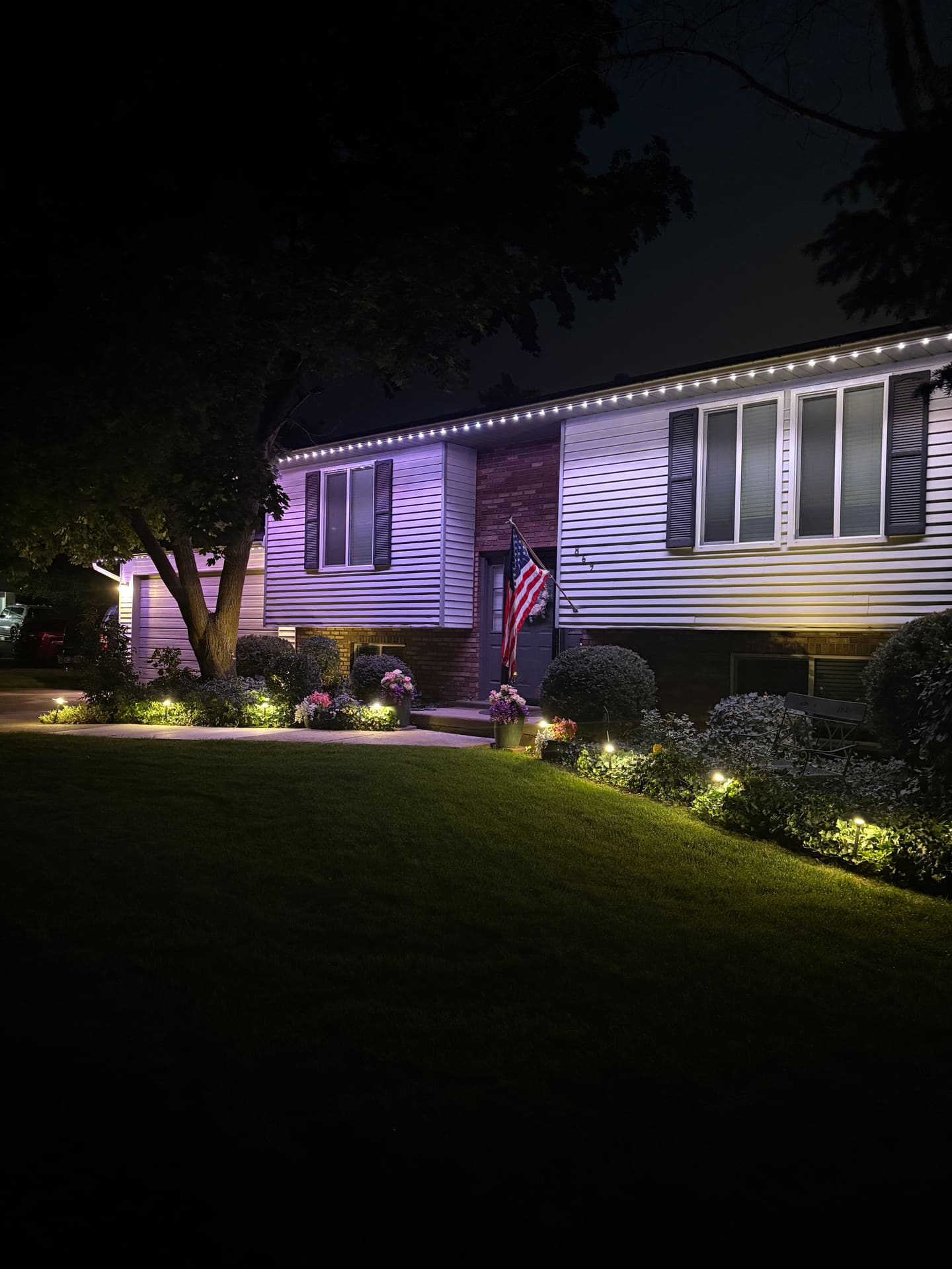 Split-level home lit purple with warm landscape lighting and an American flag