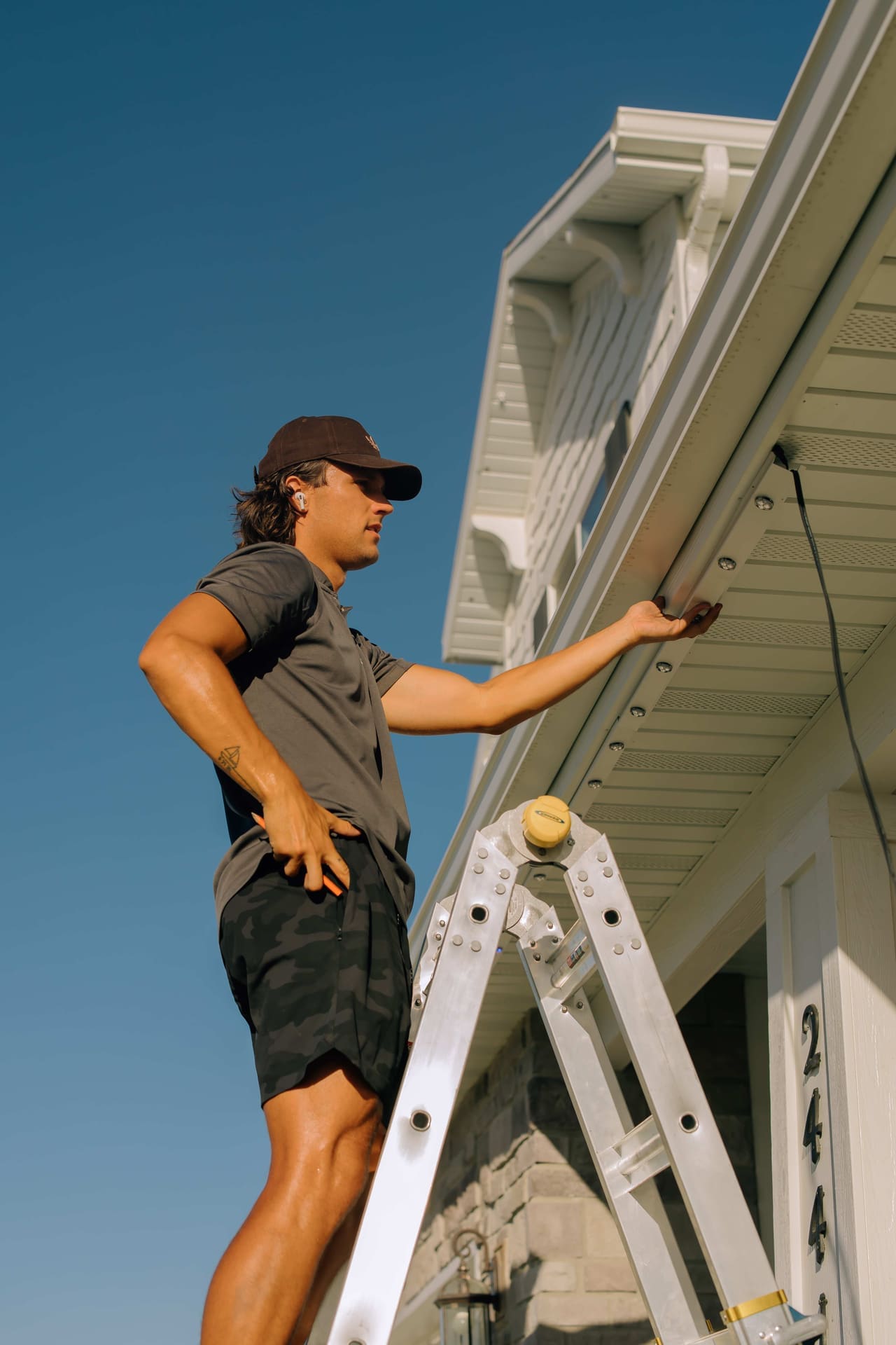 A Chipr Lux installer on a ladder fitting permanent lighting track along the eaves of a home against a clear blue sky