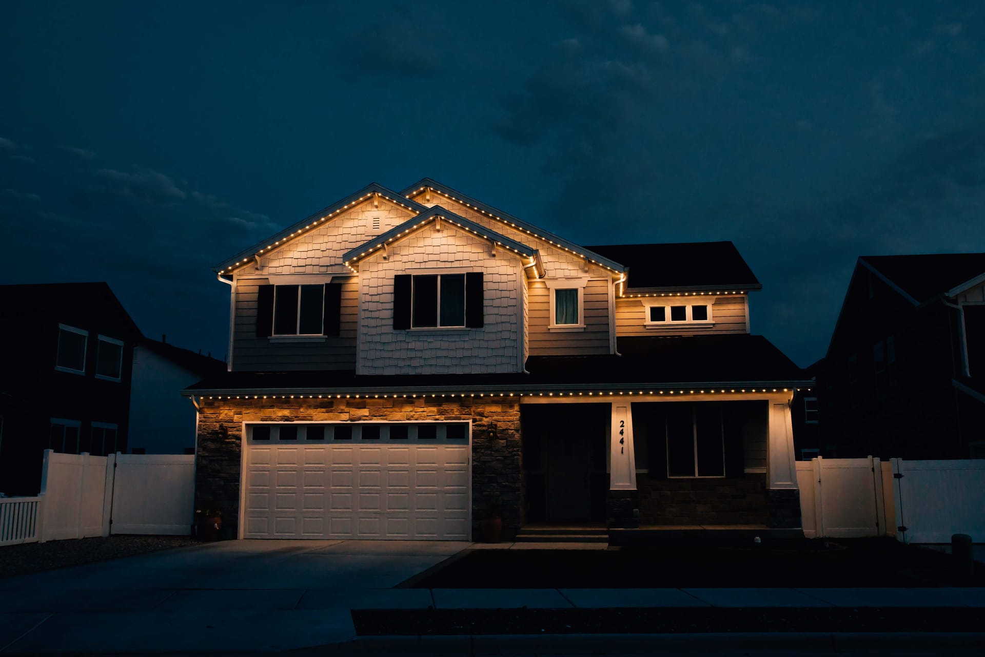 Two-story home at night with warm white permanent lights outlining the gables, rooflines, and porch